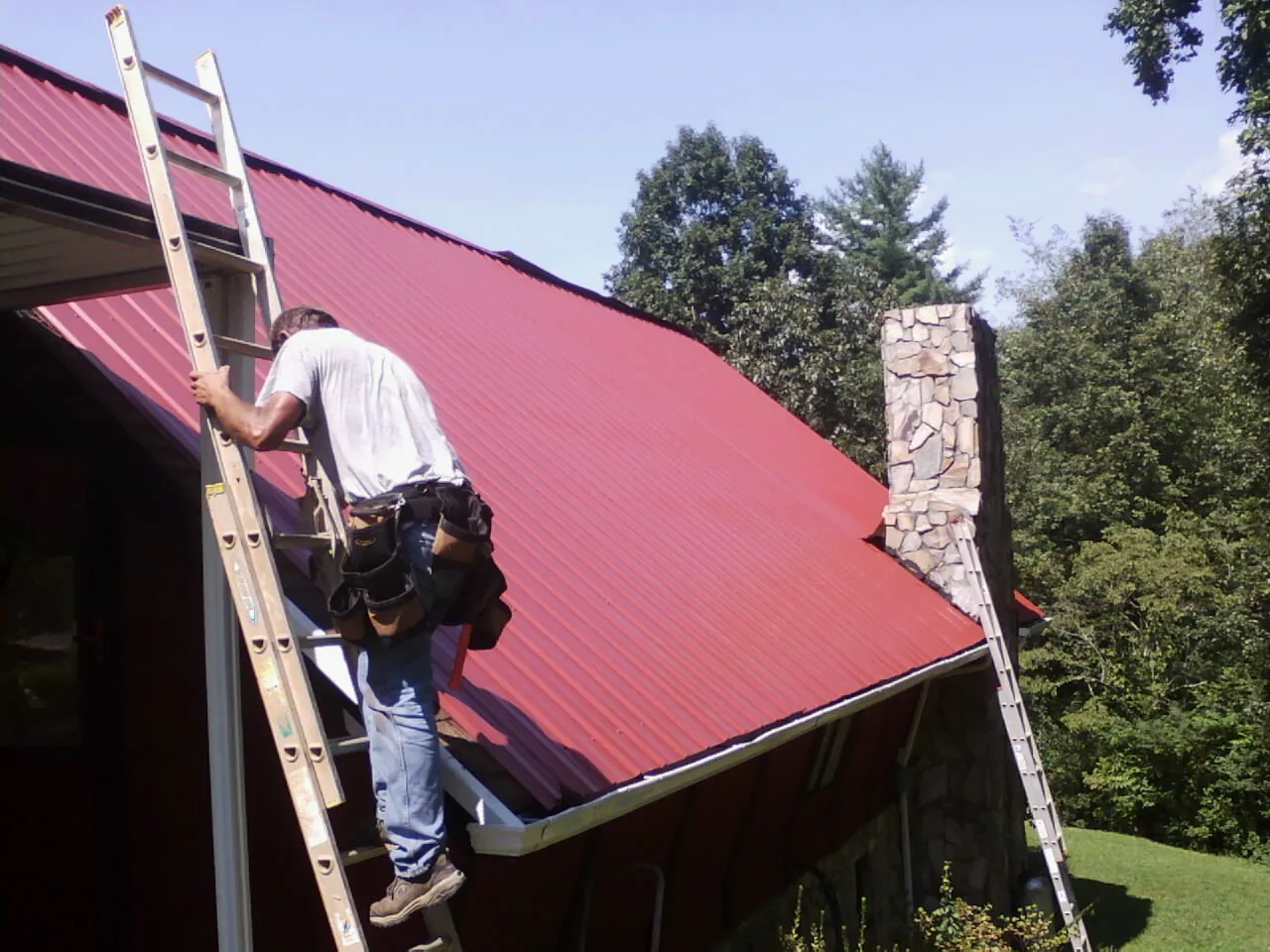 Roofer on a ladder beside a red metal roof and stone chimney — Soffit & Fascia Repair by Roofing Brothers in Campus